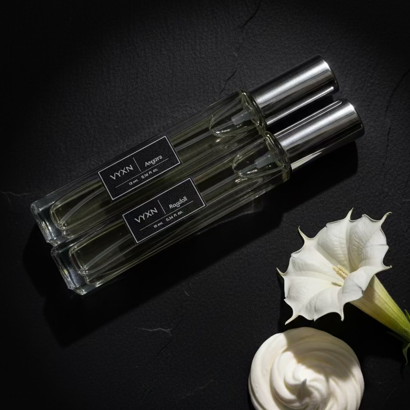 Two clear glass perfume bottles with silver caps and black labels reading VYXN, one labeled Angora and the other Ragdoll, resting on a reflective surface inside an ornate gold mirror.