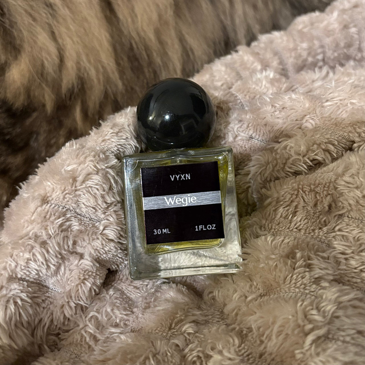 A 30 ml glass perfume bottle with a black spherical cap and a black label reading 'VYXN Wegie', resting on a fluffy beige blanket.