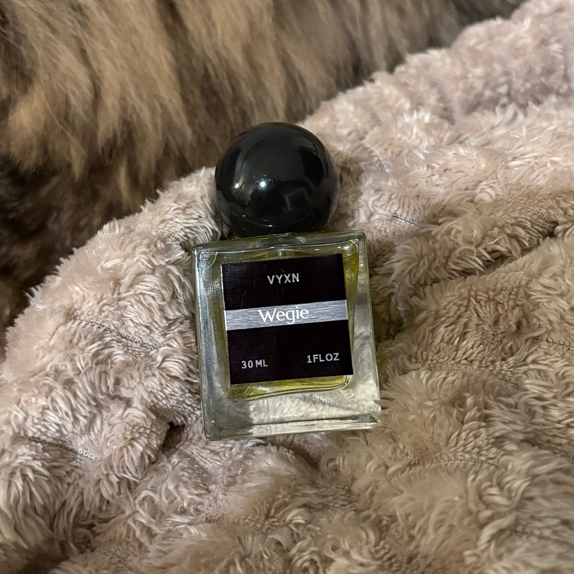 A 30 ml glass perfume bottle with a black spherical cap and a black label reading 'VYXN Wegie', resting on a fluffy beige blanket.