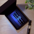 Two perfume samples in a black box on a wooden surface and one perfume sample next to the black boxwith a plant in the corner.