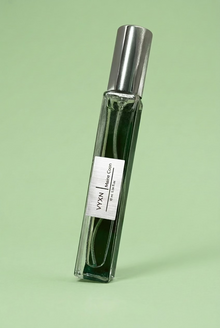 A clear rectangular glass roller bottle with a silver cap, containing dark green liquid, standing on a shiny lavender satin fabric background. The label reads 'VYXN / MAINE COON'.