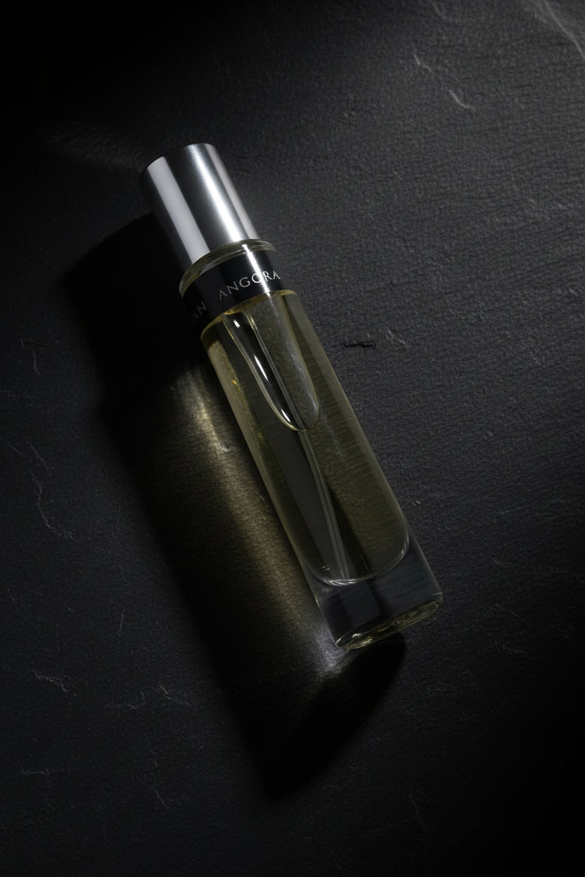 A clear glass perfume roller bottle filled with pale yellow liquid, featuring a silver cap and a black label with white text, resting on a reflective surface next to an ornate bronze mirror frame.