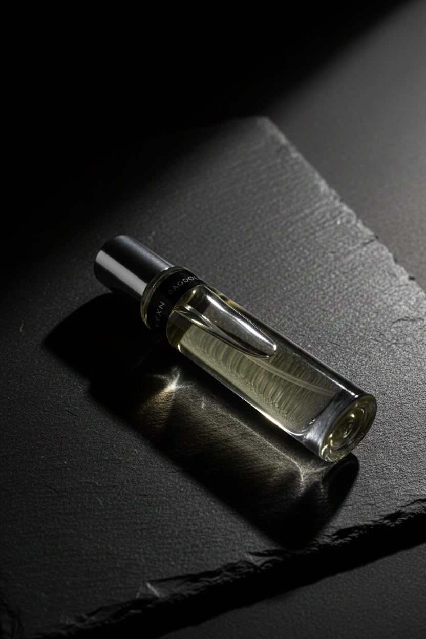 A clear glass perfume bottle with pale yellow liquid and a silver cap, resting on a reflective surface near an ornate bronze mirror frame. The label shows the brand name 'RAGDOU' partially visible.