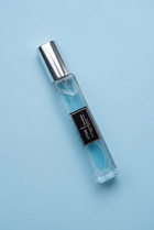 A clear rectangular perfume bottle filled with light blue liquid, featuring a black label with white text and a reflective silver cap, set against a draped lavender satin fabric.