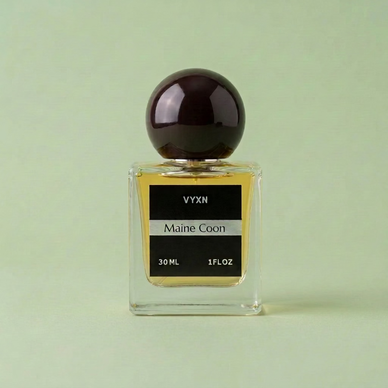 A 30 ml bottle of VYXN Maine Coon fragrance, featuring a clear glass bottle with yellow-amber liquid, a black label, and a dark marbled spherical cap, set against a draped light pink satin fabric background.
