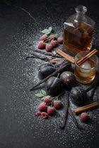 whiskey, plums, raspberries, cinnamon, vanilla pods, whiskey glass, cinnamon in whiskey glass with black slate background and dusted snow. 